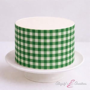 May include: A round cake with white frosting and a green and white checkered pattern around the sides. The cake sits on a white, pedestal cake stand. The cake is a dessert item.