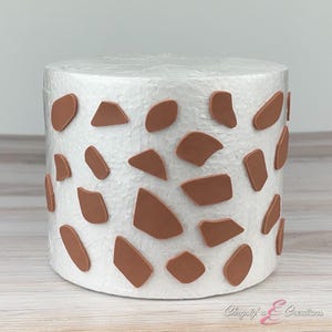 May include: A cylindrical, white decorative item with a textured surface. Brown, irregularly shaped accents are scattered across the surface. The item is likely a decorative piece for a home or event.