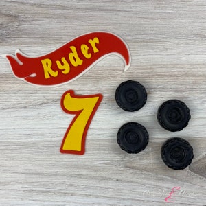 K&ouml;nnte beinhalten: Ein rotes und gelbes Banner mit dem Namen Ryder darauf. Eine gelbe Zahl 7 und vier schwarze Reifen-f&ouml;rmige Tortendekorationen.