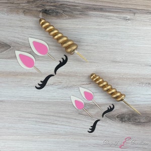 May include: A set of unicorn cake toppers, including a gold horn, pink ears, and black eyelashes. The toppers are on a wooden surface.