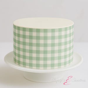 May include: A round cake with a light cream-colored top and a green and white gingham pattern around the sides. The cake sits on a white pedestal cake stand. The overall design is simple and elegant.