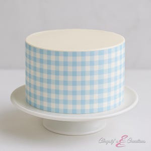 May include: A round cake with a light blue and white gingham pattern on the sides, topped with a smooth, off-white frosting. The cake sits on a white, pedestal cake stand. The cake is a dessert item.