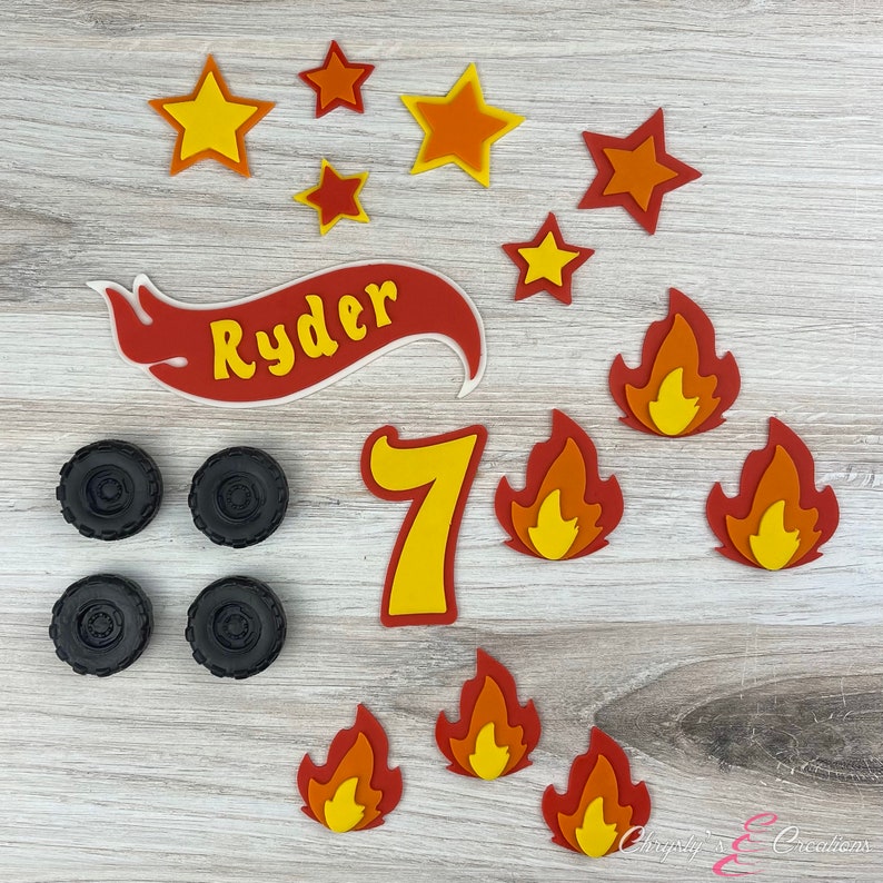 K&ouml;nnte beinhalten: Ein Set aus roten, orangen und gelben Filz-Kuchen-Toppern mit dem Namen "Ryder" und der Zahl "7" in einem roten Banner, Sternen und Flammen.