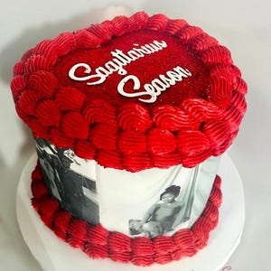 May include: A round cake with red frosting and the words "Sagittarius Season" in white. The cake has a white base with black and white photos wrapped around it. The cake is on a white cake board.