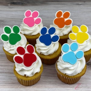May include: Seven cupcakes with white frosting, each topped with a colorful paw print. The paw prints are in shades of pink, orange, green, blue, yellow, red, and light blue. The cupcakes sit on a light wood-grain surface.