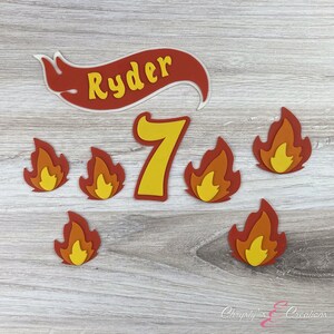 K&ouml;nnte beinhalten: Ein Set aus roten und gelben Tortendekorationen mit Flammenmotiv, der Zahl 7 und einem Banner mit dem Namen "Ryder".