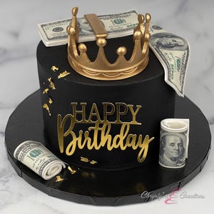 May include: A black birthday cake decorated with a gold crown, gold lettering that says "Happy Birthday", and edible dollar bills. The cake sits on a black cake board.