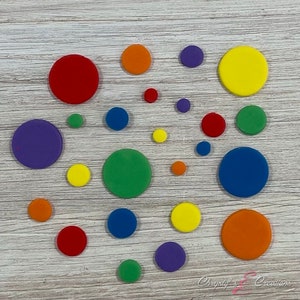 May include: A collection of colorful, flat, circular objects in various sizes and shades, including red, orange, yellow, green, blue, and purple. The circles are arranged on a light wood-grain surface.