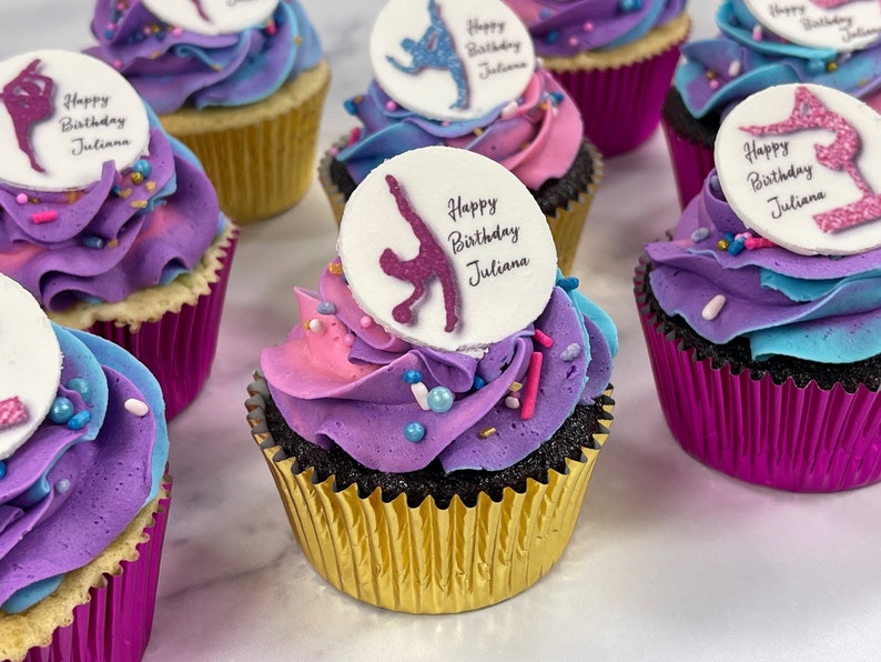 May include: A close-up of cupcakes decorated with purple, pink, and blue frosting. Each cupcake has a round edible image on top with a gymnast silhouette and the text "Happy Birthday Juliana".