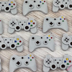 May include: A collection of light gray video game controller-shaped decorations. Each controller has colorful buttons and a directional pad. The controllers are arranged on a light wood-grain surface.