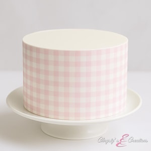 May include: A round cake with a light pink and white gingham pattern on the sides, sitting on a white cake stand. The top of the cake is a solid off-white color. The cake stand has a pedestal base.