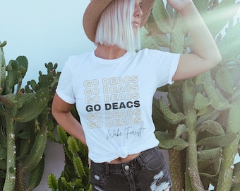 May include: White t-shirt with the text "GO DEACS" repeated in a stacked pattern, with one line in black. The words "Wake Forest" are written in script below the stacked text. The shirt is worn with black denim shorts.