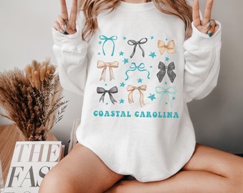 May include: White sweatshirt with a graphic design of blue, gray, and tan bows with stars. The text "Coastal Carolina" is printed in blue below the bows.