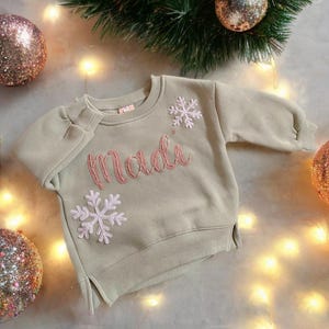 Könnte beinhalten: Ein hellgrüner Pullover mit dem Namen "Madi", der in rosafarbenem Garn gestickt ist. Der Pullover hat zwei weiße Schneeflocken-Applikationen.
