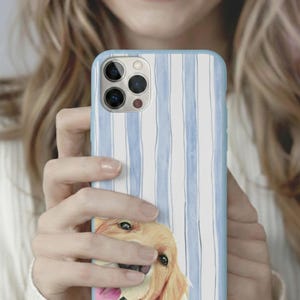 May include: A light blue and white striped phone case featuring a watercolor illustration of a golden retriever dog. The dog's face is visible at the bottom of the case, with its tongue sticking out. The case is held by a person.