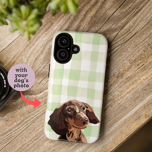 May include: A phone case featuring a dachshund illustration. The case has a light green and white gingham pattern and the text "with your dog's photo". The dog illustration is brown and tan. The phone case is on a wooden surface.