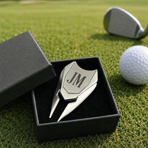 May include: A silver-toned metal golf divot tool featuring a shield-shaped top engraved with the letters JM. The tool is presented inside a black box with a soft black interior lining, showcasing a sleek, polished finish and a two-pronged design.
