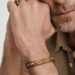 May include: A link bracelet constructed from alternating dark and light brown wood segments. The central rectangular piece features the engraved text "LOGAN PIERCE" flanked by two small star symbols. The bracelet has a smooth, polished finish.