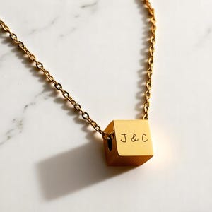 May include: A gold-toned necklace featuring a small, smooth cube pendant suspended from a delicate chain. The front face of the cube is engraved with the letters J & C in a simple, clean font. The metal has a polished finish that reflects light.