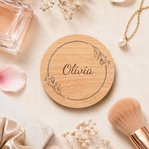 May include: A circular wooden disc featuring the name Olivia engraved in the center, surrounded by a thin circular border with delicate floral line art accents on the left and right sides of the frame.
