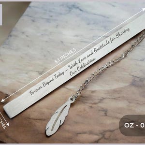 May include: Silver bookmark with a feather charm and the inscription "Forever Begins Today - With Love and Gratitude for Sharing Our Celebration." The bookmark is 5.1 inches long and 0.4 inches wide and weighs 0.95 ounces.