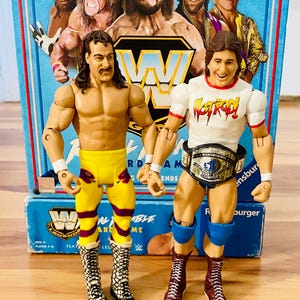 May include: Two action figures of wrestlers stand in front of a blue box featuring images of other wrestlers. One figure wears yellow pants with snake designs and boots. The other figure wears a white shirt with "Hot Rod" text and a championship belt.