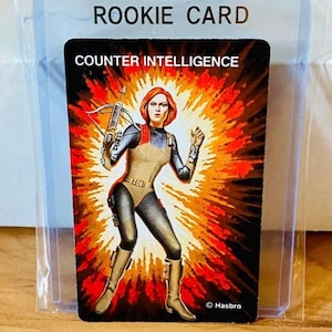 May include: A trading card featuring a female character with red hair, holding a crossbow, and wearing a tan and black bodysuit. The card is labeled "ROOKIE CARD" and "COUNTER INTELLIGENCE" with a fiery background.