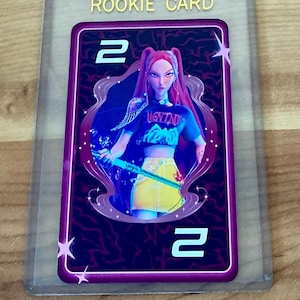 May include: A rectangular trading card in a clear plastic sleeve featuring the text ROOKIE CARD at the top. The card has a dark purple background with a lightning pattern, the number 2 in the corners, and an illustration of a character with long red hair wearing a black crop top and yellow skirt holding a glowing blue staff.