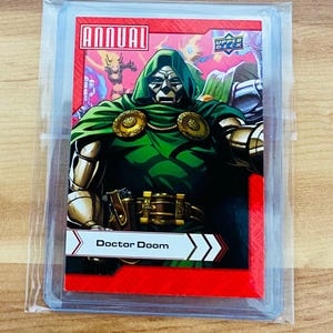 May include: A Doctor Doom trading card from the Upper Deck Annual series, encased in a protective sleeve. The card features a vibrant illustration of Doctor Doom in a green hooded cloak and metallic armor, set against a red background. The card is in a clear plastic sleeve.