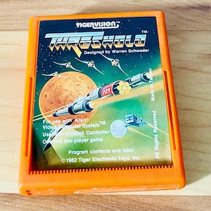 May include: An orange Atari video game cartridge for the game "Threshold." The label features a space scene with a spaceship, planets, and the game's title. The cartridge is labeled "Made in Taiwan."