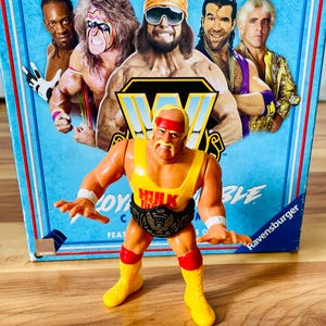 May include: A plastic Hulk Hogan action figure wearing a yellow tank top with red text, red wristbands, red knee pads, and yellow boots, featuring a black championship belt. The figure stands before a blue board game box featuring WWE wrestlers.