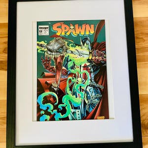 May include: A framed comic book titled Spawn issue 15, featuring a colorful illustration of a character with a skull-like face and green energy effects. The comic is encased in a black frame with a white mat board, displaying the Image logo.
