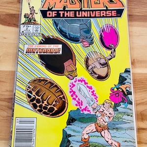 May include: A vintage "Masters of the Universe" comic book. The cover features He-Man wielding a sword, with the title text in orange and black. The background is yellow with images of the Meteorb robots. The comic is from July.