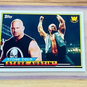 May include: A Topps trading card featuring Stone Cold Steve Austin. The card shows two images of the wrestler, one with his arms raised. The card is in a protective sleeve. The text "STONE COLD STEVE AUSTIN" is visible.