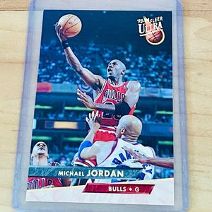 May include: A 1993 Fleer Ultra basketball card featuring Michael Jordan in a red Bulls uniform, mid-jump, with the ball in his hand. The card is in a protective sleeve. The card also has the text "Bulls G".