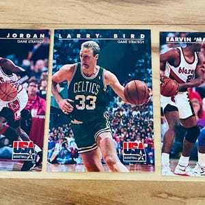 May include: Three basketball trading cards featuring Michael Jordan, Larry Bird, and Earvin 'Magic' Johnson. Each card displays a player in action with a basketball, with the USA Basketball logo at the bottom.