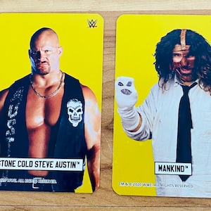 May include: Two yellow trading cards featuring WWE wrestlers. One card shows Stone Cold Steve Austin in a black vest with a skull emblem. The other card features Mankind in a white shirt and tie, holding a glove with a face.