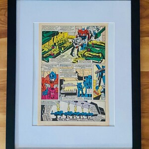 May include: Framed comic book art featuring Transformers characters in action. The artwork is in a black frame with a white mat. The comic panels show robots in red, blue, and white, set against a backdrop of yellow and green machinery.