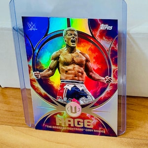 May include: A Topps trading card featuring a wrestler in a dynamic pose, set against a vibrant background of red and blue hues. The card is labeled "RAGE" with the text "The American Nightmare" Cody Rhodes. The card is encased in a protective sleeve.