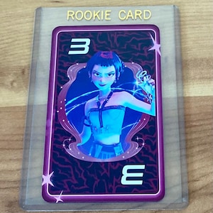 May include: A collectible trading card encased in a clear plastic protector labeled ROOKIE CARD. The card features a dark purple background with a stylized illustration of a person with short dark hair wearing a blue top, framed by a decorative border.
