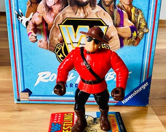 May include: A red and black action figure of a wrestler stands on a WWF Wrestling card in front of a WWF board game box. The box features images of several wrestlers. The game is made by Ravensburger.