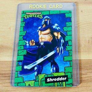 May include: A Teenage Mutant Ninja Turtles "Rookie Card" featuring Shredder. The card has a green border with a brick wall design. Shredder is depicted in a fighting pose, holding a sword. The card is encased in a clear protective sleeve.