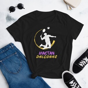 May include: Black t-shirt featuring a white badminton player silhouette within a yellow circle, with a coffee cup graphic. The text "MACTAN DALGONAS" is in purple and yellow. The shirt is paired with blue jeans and black sneakers.