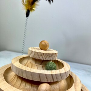 May include: A wooden cat toy with three tiers of circular tracks. The toy has four balls, two brown and two green, that roll around the tracks. A feather teaser is in the background.