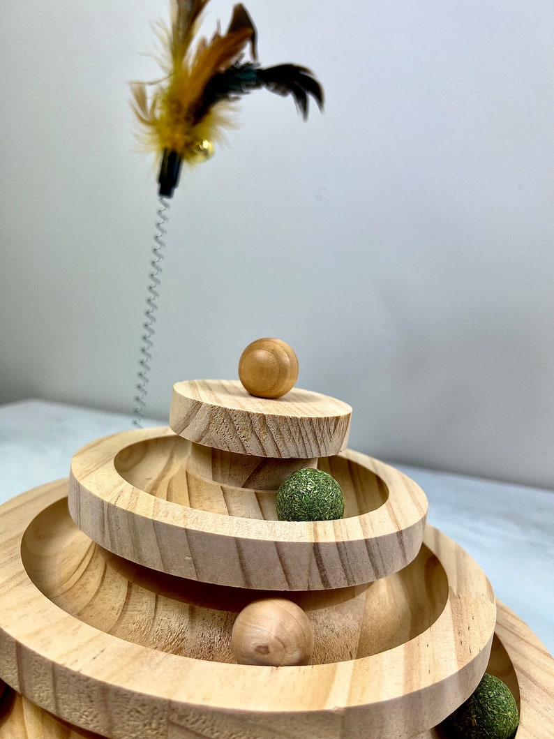 May include: A wooden cat toy with three tiers of circular tracks. The toy has four balls, two brown and two green, that roll around the tracks. A feather teaser is in the background.