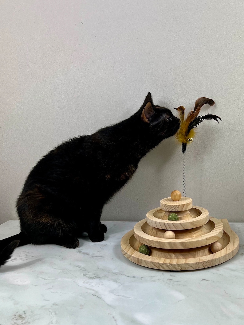 May include: A black cat playing with a wooden cat toy with a feather teaser. The toy has three tiers with rolling balls.
