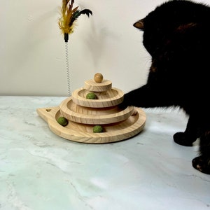 May include: A black cat plays with a wooden cat toy with green balls and a feather teaser. The toy has three tiers and is on a white marble surface.