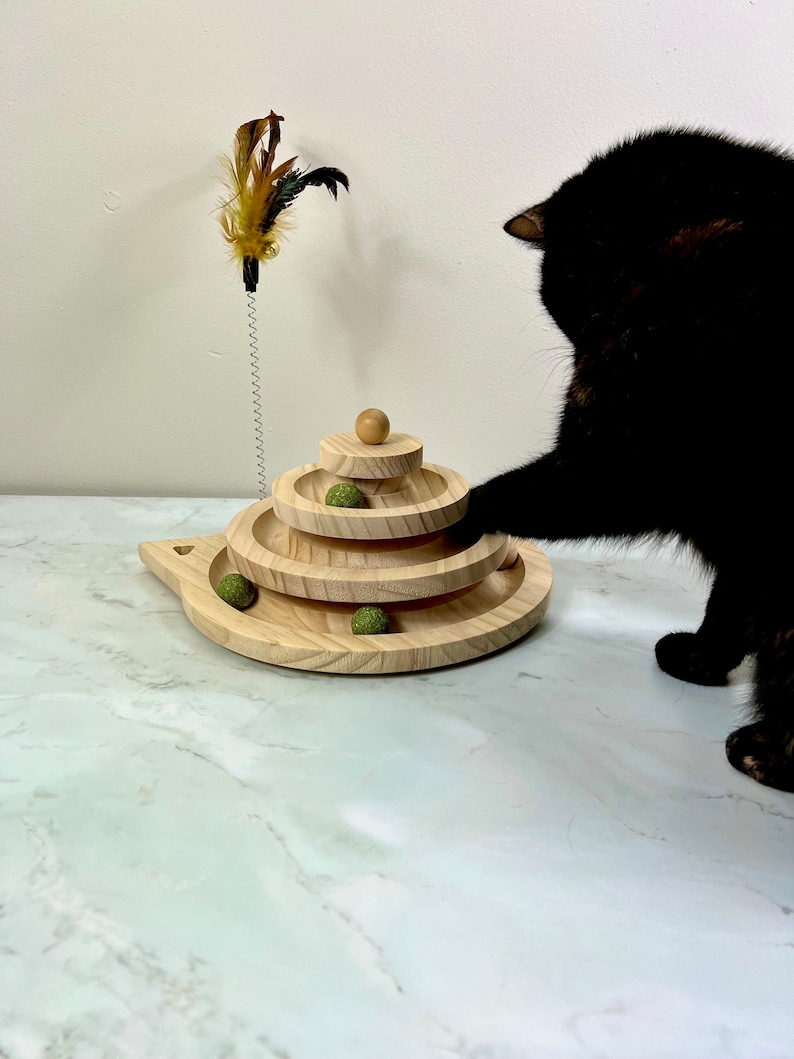 May include: A black cat plays with a wooden cat toy with green balls and a feather teaser. The toy has three tiers and is on a white marble surface.