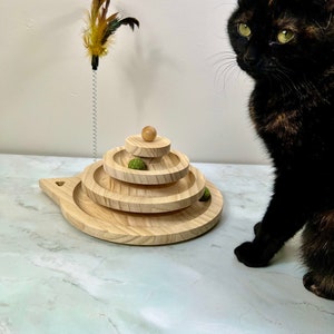 May include: A wooden cat toy with three tiers and a small wooden ball on top. The toy is made of natural wood and has a light brown color. A black cat is sitting next to the toy, looking at it.
