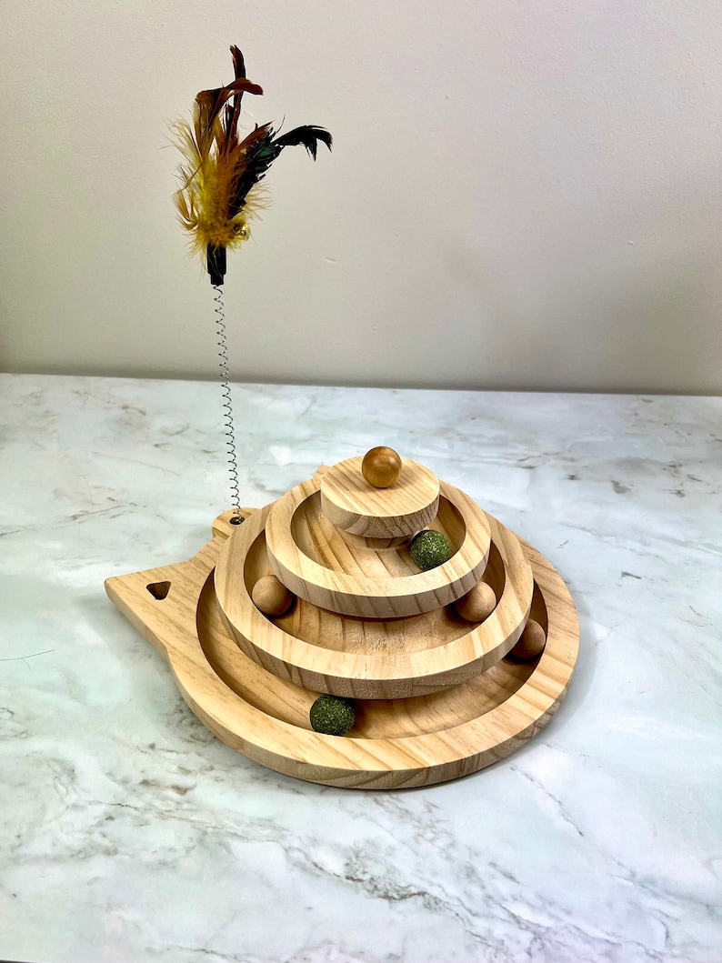 May include: Wooden cat toy with a spring-loaded feather teaser and a multi-level track with rolling balls. The toy is shaped like a cat's head and has a natural wood finish.